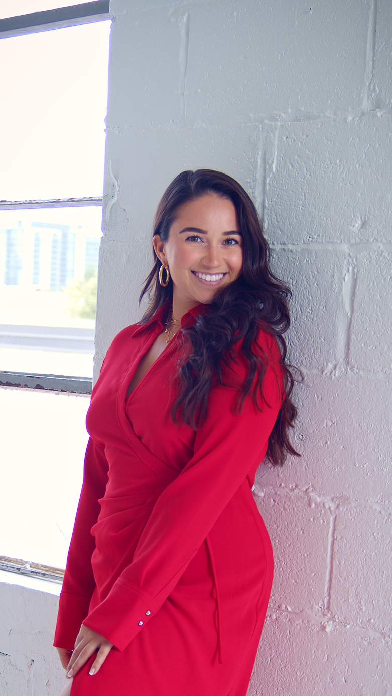 Young woman with brown wavy hair in bright red dress leaning on white brick wall